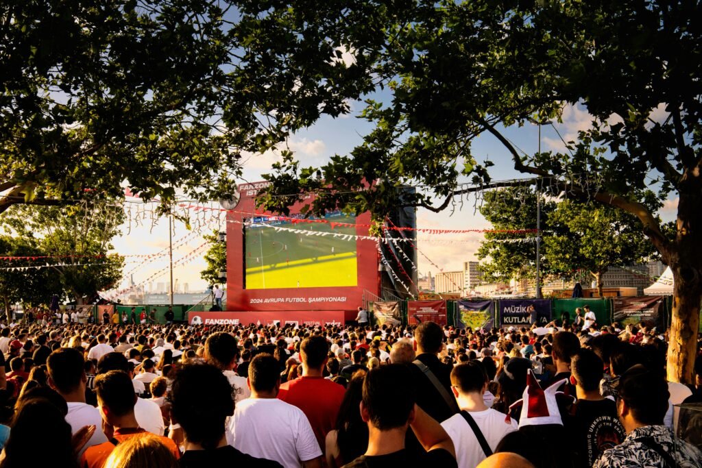Crowd gathered in İstanbul park watching Euro football on big screen. Energetic summer atmosphere.